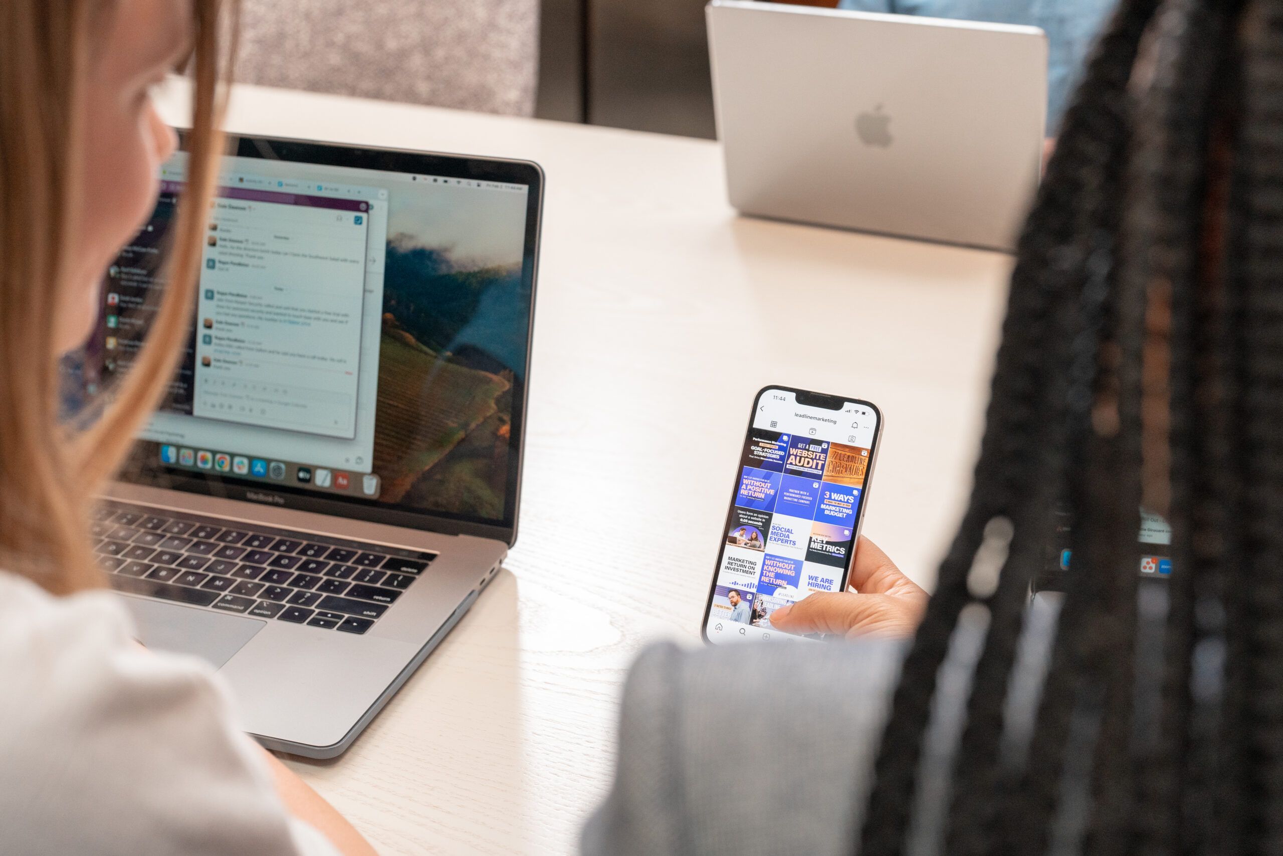 Two teammates review results on multiple devices—a Slack thread on a laptop and performance posts on a phone—underscoring revenue accountability.