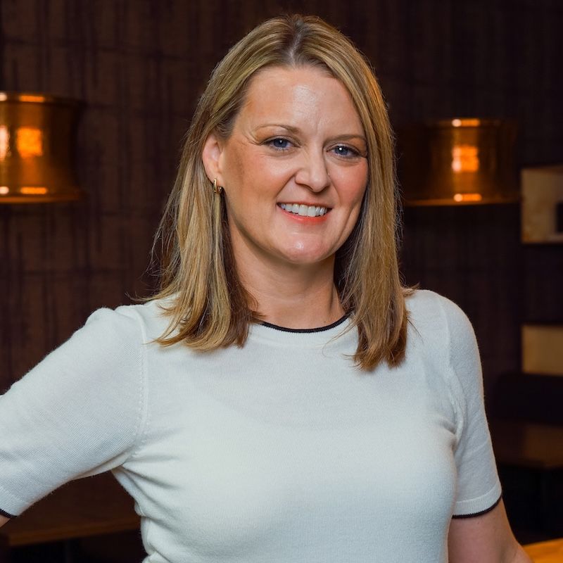 A professional headshot of a woman smiling confidently, wearing a light short-sleeve top, standing indoors with warm lighting in the background.