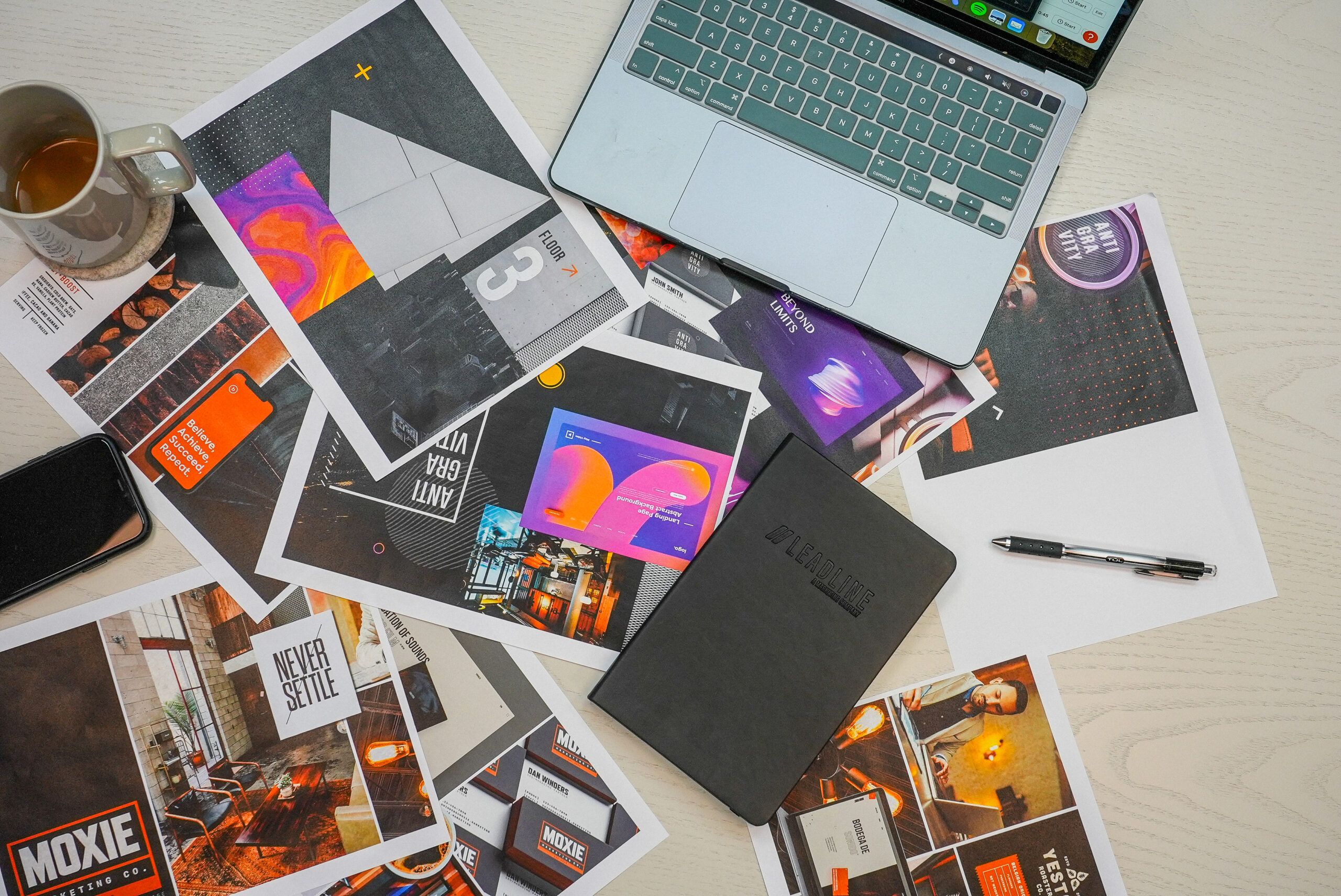 Overhead view of a busy desk: a laptop, mug of tea, phone, pen, and black notebook sit amid scattered printouts of vibrant ads and mood-board images, conveying nonstop creative iteration.
