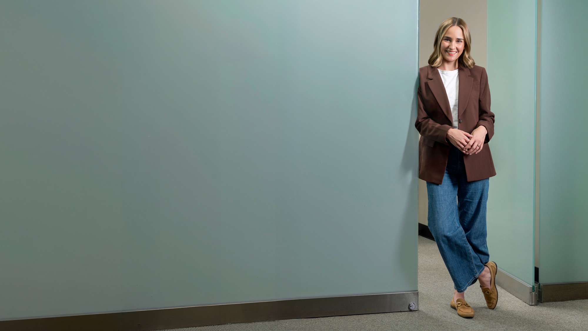 Smiling professional in a brown blazer beside a frosted-glass office wall
